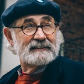 Older man with round glasses, a blue hat and a neatly trimmed salt and pepper beard